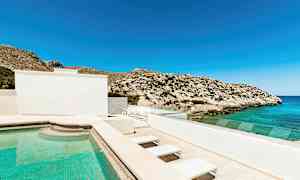 Et utendørsbasseng ved et hus, med utsikt over havet og steinete fjell under en klarblå himmel. (AI-generert tekst)