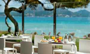 Bildet viser en terrasse med hvite bord og stoler og en utsikt over blått hav og grøntområder. (AI-generert tekst)