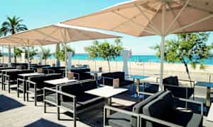 En utendørskafé med bord, stoler og parasoller, med utsikt over en sandstrand og havet. (AI-generert tekst)