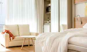 Et lyst hotellrom med en sofa, et lite bord og en oppredd seng. I bakgrunnen er det et minikjøkken med kjøleskap. (AI-generert tekst)