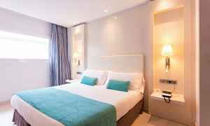 Et lyst hotellrom med en dobbeltseng, to turkise puter, et teppe og vegglamper på hver side av sengen. (AI-generert tekst)