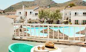 Utsikt fra en balkong mot et basseng og bygninger med fjell i bakgrunnen. (AI-generert tekst)