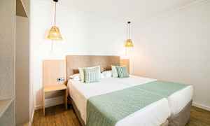 Et hotellrom med to oppredde enkeltsenger, grønne sengetepper og puter samt to hengende lamper. (AI-generert tekst)