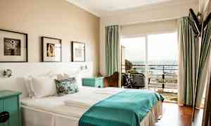 Et hotellrom med en dobbeltseng og sengetøy i hvitt og blått, veggkunst over sengen og en balkong med utsikt over havet. (AI-generert tekst)