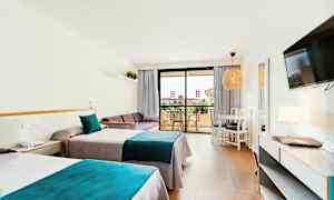 Et hotellrom med to enkeltsenger med blå og grå sengetøy, en sofa, et bord med stoler og en balkong i bakgrunnen. (AI-generert tekst)