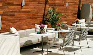 Et stilrent møblement på en terrasse med bord og stoler, dekorert med planter og puter, mot en trepanelvegg. (AI-generert tekst)