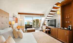 Et hotellrom med tremøbler, en hvit seng, en trapp og utsikt mot en terrasse med hav i bakgrunnen. (AI-generert tekst)