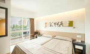 Et hotellrom med to enkeltsenger, dekorert med mønstrede sengetepper. Det er en møblert balkong som vender ut mot uteområdet. (AI-generert tekst)