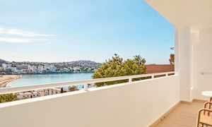 Utsikt fra en balkong over en strand ved en bukt, med bygninger og grøntområder i bakgrunnen. (AI-generert tekst)