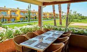 Bilde av en uterrasse med et dekket bord og stoler, med utsikt over en hage og gule bygninger i bakgrunnen. (AI-generert tekst)