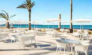 Utendørs sitteområde med hvite møbler og parasoller, samt palmer i bakgrunnen ved sjøen. (AI-generert tekst)