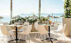 Bilde av en uteservering med hvite stoler og bord, med utsikt over havet og parasoller i bakgrunnen. (AI-generert tekst)