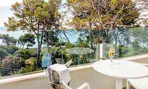 En balkong med utsikt over trær og havet. På balkongen står et hvitt bord med et glass champagne og en bøtteholder med en champagneflaske. (AI-generert tekst)