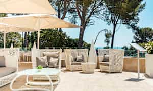 Sitteområde utendørs med hvite flettede møbler, beige puter og parasoller. Grønt landskap og sjøen i bakgrunnen. (AI-generert tekst)