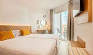 Et lyst hotellrom med en stor seng, to gule puter, en skrivebordshjørne og en balkong med utsikt. (AI-generert tekst)