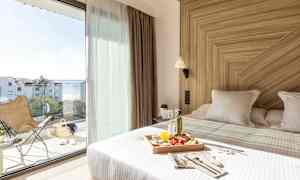 Et lyst hotellrom med en oppredd seng med en frokostbrett. Store glassdører fører ut til en balkong med utsikt over havet. (AI-generert tekst)