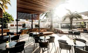 En utendørsterrasse med sitteplasser, bord og solbeskyttelse under et treskjermtak. Palmer og parasoller er synlige i bakgrunnen. (AI-generert tekst)