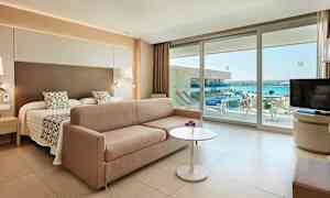 Hotellrom med en seng, sofa, bord og utsikt over havet gjennom glassdører. (AI-generert tekst)