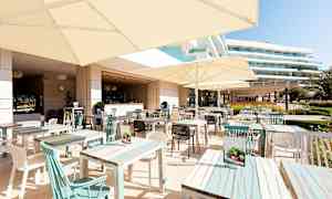 En utendørsrestaurant med bord og parasoller, omkringliggende bygninger er synlige i bakgrunnen. (AI-generert tekst)