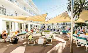 En utendørsterrasse ved en bygning, med bord, stoler og skyggefulle markiser. (AI-generert tekst)