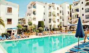 Et bassengområde med solsenger rundt et klart blått basseng, foran en hvit bygning med balkonger. (AI-generert tekst)