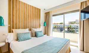 Et moderne hotellrom med en stor seng, trepanel bak sengen, hvite og blå sengetøy, samt en vindusutsikt over bygninger og trær utenfor. (AI-generert tekst)