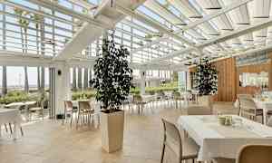 Restaurant med bord og stoler under et glasstak, omgitt av grønne planter og palmer. (AI-generert tekst)
