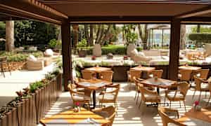 En åpen restaurant med bord og stoler, omgitt av grøntområder og utsikt mot en hage og basseng. (AI-generert tekst)