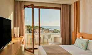 Et hotellrom med en seng, skrivebord, TV og en åpen balkongdør som leder til en balkong med utsikt over havet og trær. (AI-generert tekst)