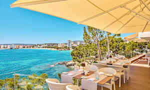 Uteservering med utsikt over en klarblå havsbukt og bygninger i det fjerne. (AI-generert tekst)