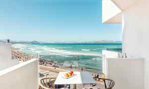 Utsikt fra en balkong mot en strand med turkist vann, sand og mennesker som bader. (AI-generert tekst)