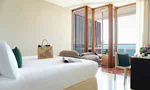 Et hotellrom med en seng, skrivebord med stol, og en balkong med utsikt over havet. (AI-generert tekst)