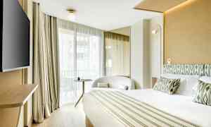 Et hotellrom med en stor seng, lenestol, bord og en TV, samt store vinduer med lyse gardiner. (AI-generert tekst)