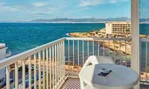 Utsikt fra en balkong mot en kyst med hav og bygninger i bakgrunnen. (AI-generert tekst)