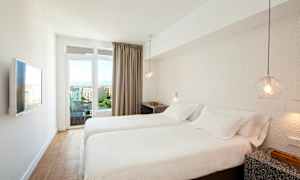 Et hotellrom med to enkeltsenger, hvitt sengetøy, en vegg-TV og utsikt over en by gjennom glassdører. (AI-generert tekst)