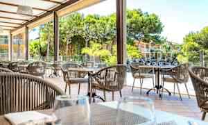 Utendørs sitteområde med bord og stoler, omgitt av grønne trær og en bygning i bakgrunnen. (AI-generert tekst)