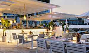 En utendørsrestaurant med hvite møbler og grønne planter, som ligger nær en bygning med glassvinduer. (AI-generert tekst)