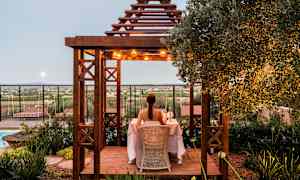 En person sitter ved et bord under en trepergola dekorert med lys, omgitt av grønne omgivelser med utsikt over landskapet. (AI-generert tekst)