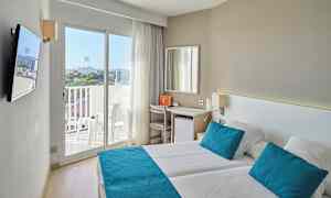 Et lyst hotellrom med to senger, blå puter og sengetepper, samt en balkong med utsikt. (AI-generert tekst)