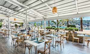 Uteservering ved sjøen med bord og stoler samt utsikt over båter i havnen. (AI-generert tekst)