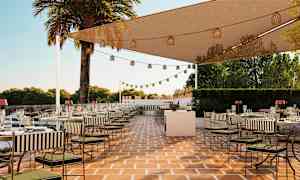 En utendørsterrasse med bord og stoler, pyntet med lyslenker under et beskyttende nettak. I bakgrunnen ses trær og en klar himmel. (AI-generert tekst)