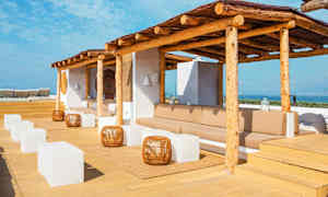 Terrasse med solbeskyttelse av tre, sittegrupper, hvite kubebord og utsikt over havet. (AI-generert tekst)