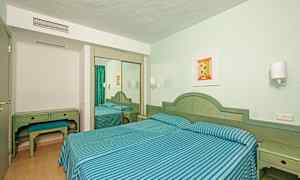 Et hotellrom med to enkeltsenger dekket med blå- og grønnstripete sengetøy, skrivebord med krakk og et speilskap. (AI-generert tekst)