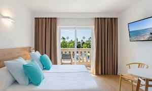Et hotellrom med to enkeltsenger med lyseblå og turkise puter, en balkong med utsikt over hagen, og en TV på veggen. (AI-generert tekst)