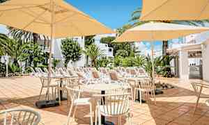 Bilde av en terrasse med bord, stoler og parasoller i en solrik setting. I bakgrunnen er det solsenger og grøntområder. (AI-generert tekst)