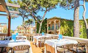 Bilde av en utendørs terrasse med dekkede bord og stoler, omgitt av trær og bygninger med plantekledde fasader. (AI-generert tekst)