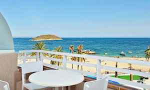 Utsikt fra en balkong med et hvitt bord og to stoler mot en sandstrand, palmer og et blått hav. (AI-generert tekst)