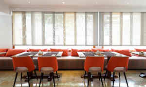 Et rom med bord og stoler, dekorert med oransje puter og solbeskyttede vinduer. (AI-generert tekst)