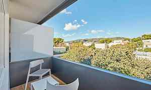 En balkong med to stoler og et bord, med utsikt over omkringliggende trær og bygninger under en blå himmel. (AI-generert tekst)