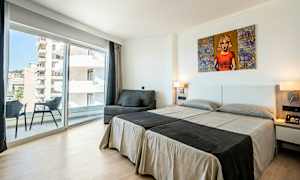 Et hotellrom med to enkeltsenger, en sofa, en balkong og store glassvinduer. (AI-generert tekst)